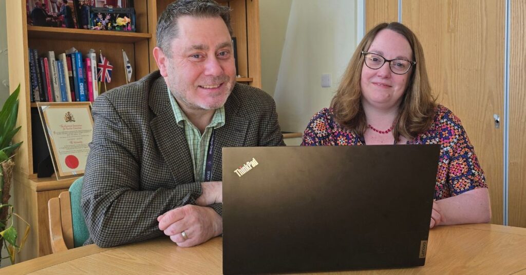 Image of a man and a woman smiling in an office room, sat behind a laptop and in front of a bookshelf